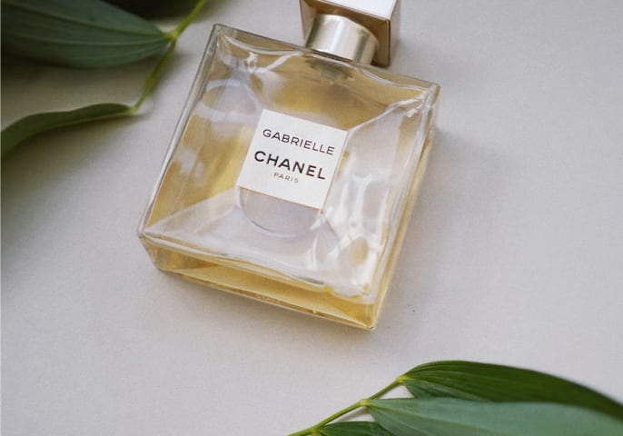 a chanel perfume bottle laid on a grey surface surrounded by green leaves